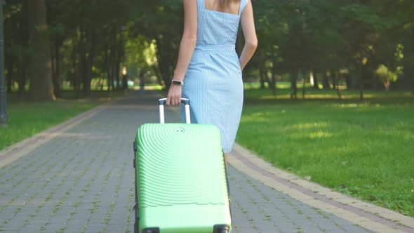 Young Woman Walking on Park Sidewalk with Green Suitcase on Summer Day