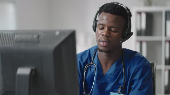 A Black Man Sits at a Computer in a Doctor's Uniform and Writes a Patient's Card While Taking Calls