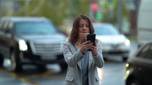 Female City Dweller with Neck Brace Is Browsing in Smartphone on Street, Walking Along Road