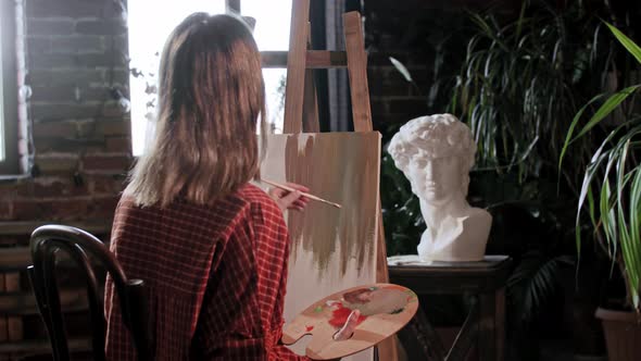 A Young Woman Artist Looking Behind the Easel and Applying Base Colors on a Canvas with a Brush