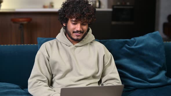 Cheerful Indian Man Wearing Casual Hoodie Sitting on the Sofa and Using Laptop at Home