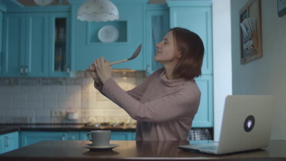 Happy woman dancing and singing into kitchenware like a microphone at home