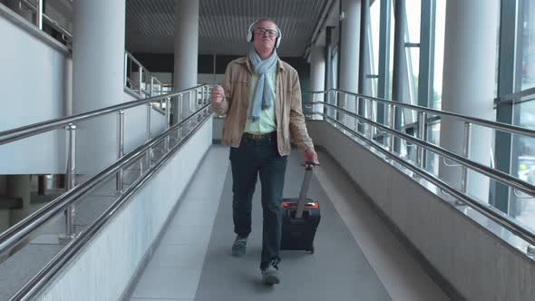 Senior Man Walking in Airport with Luggage and Listening To Music in Headphones