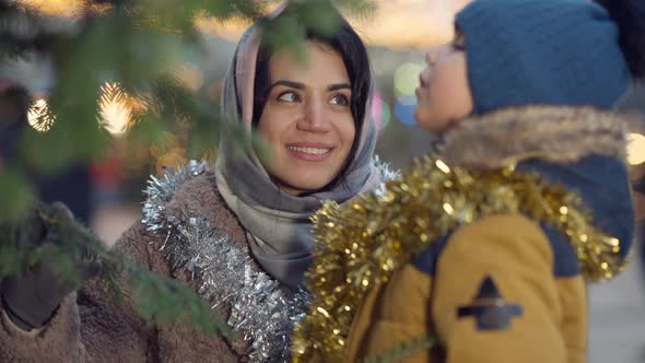 Beautiful Happy Middle Eastern Woman Admiring Decorated Christmas Tree on City Square with Boy