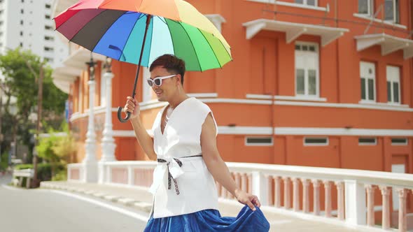 Asian lgbtq guy wearing woman clothes with sunglasses holding rainbow umbrella walking down street.