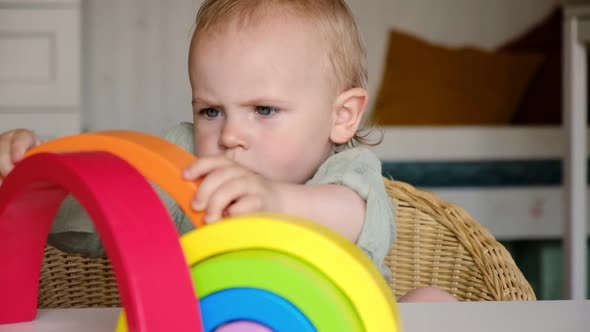 Little Baby Child in Clothes Made of Natural Fabric Plays with Rainbow Colored Wooden Toys at Table