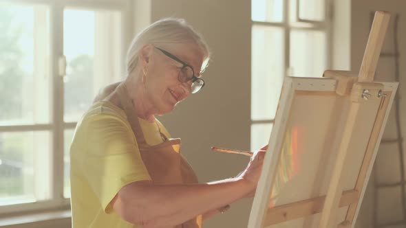 Happy Senior Woman Artist Paints a Picture on Canvas While Standing in a Sunny Workshop