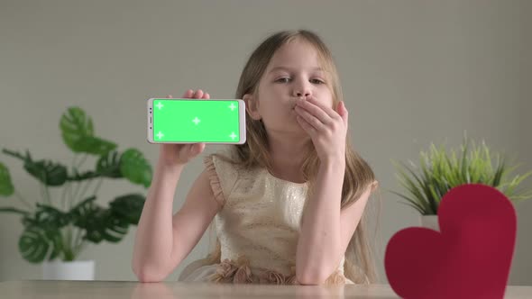 Cute Little Girl Shows Phone with Green Screen Near Red Heart and Sending Air Kiss