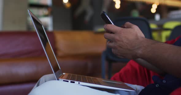 African american man wearing face mask sitting in cafe using smartphone and laptop