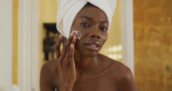 Portrait of smiling african american woman with towel using cream on her face in bathroom