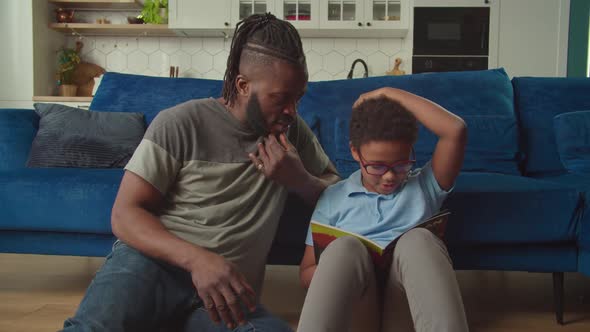 Excited African American Kid and Father Reading Comic Book on Floor at Home