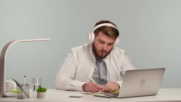 Young Man in Big White Headphones Watching Online Webinar and Making Notes Studying Remotely at Home