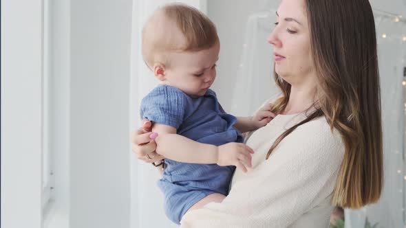 Close-up of a beautiful loving mother in a dress, who is standing at the window with a baby in her a