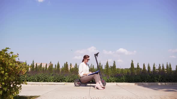 Modern Office Worker in a Cityscape. Attractive Slim Girl Is Sitting on an Electric Kick Scooter in