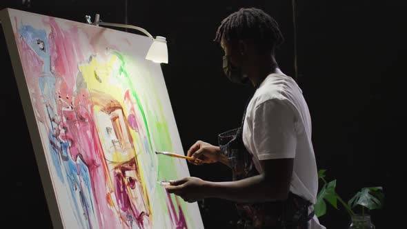 African american male artist wearing face mask painting on canvas at art studio