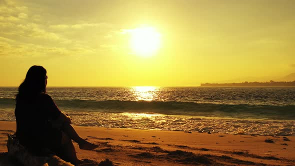 Silhouette of woman sitting on quiet beach at sunset with glowing yellow sky over troubled sea with