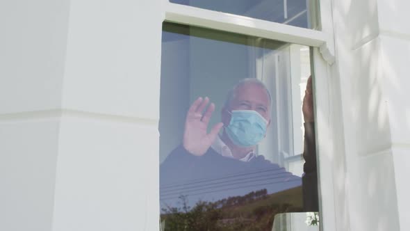 Senior caucasian man at home wearing face mask standing at a window and waving