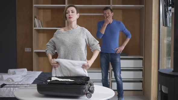 Portrait of Stressed Caucasian Attractive Woman Packing Travel Bag with Blurred Shocked Man Standing