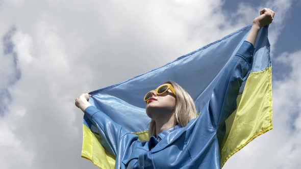 Stylish woman in blue shirt and yellow sunglasses stand with ukrainian flag on sky background