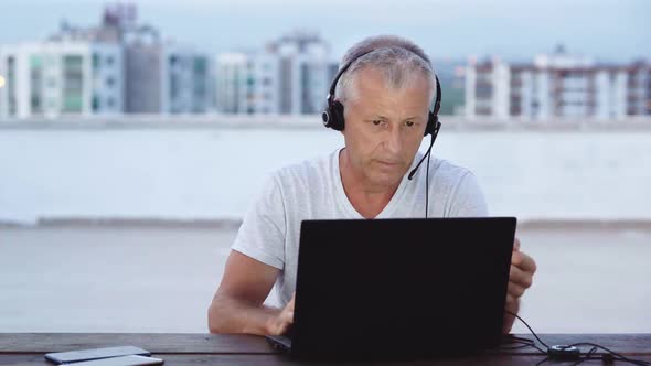 An Elderly Whitehaired Man Talks on the Internet on a Laptop with Headphones with a Microphone Late