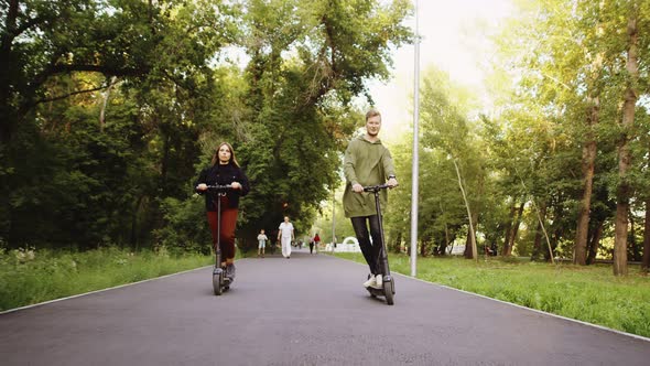 Young Couple Rides on Rented Electric Scooters Along Path in Park Front View