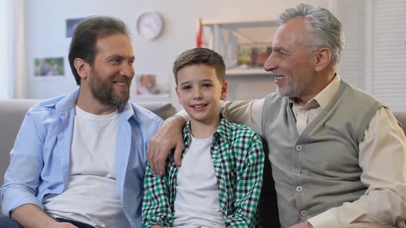 Happy Father and Grandpa Hugging Boy, Sitting on Sofa at Home, Looking at Camera