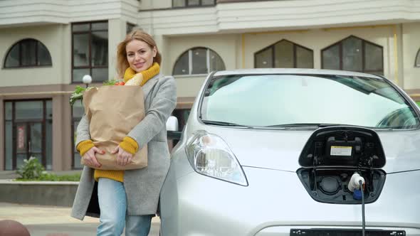 A Woman Stands Near Her Electric Car with Groceries and Looks at the Camera