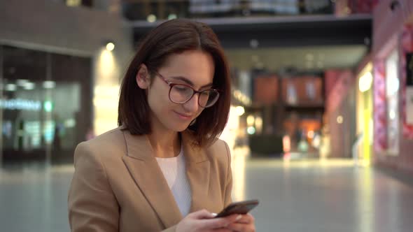 A Young Woman Sits with a Phone in Her Hands in a Shopping Center with a Large Paln. A Girl in a