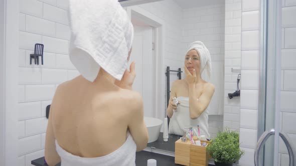 Young Woman in Towel Applying Cream To Face and Looking To Mirror at Home Bathroom. Beauty