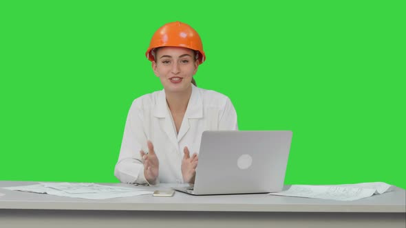 Smiling Female Engineer Talking To the Camera Sitting on the Desk with a Lot of Papers on a Green