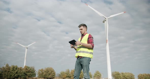 Young Engineer with Tablet in Hand Looks Behind and Works on Electric Turbines