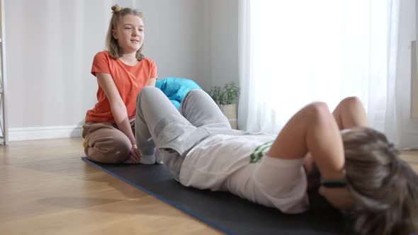 Wide Shot of Cute Positive Girl Holding Feet of Boy Pumping Press on Exercise Mat