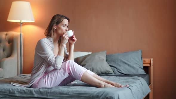 Happy Relaxed Barefoot Young Woman Drinking Hot Tea in Bedroom Enjoying Weekend at Home