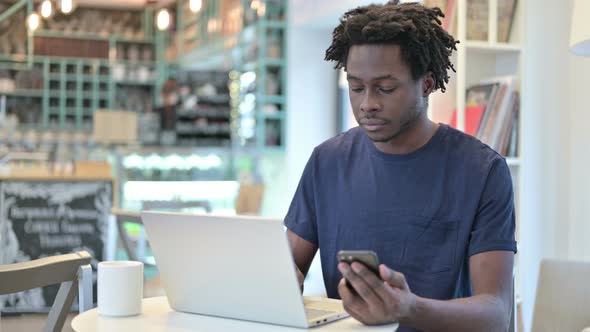 African Man Using Smartphone and Laptop in Cafe