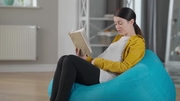 Side View Smiling Pregnant Woman Reading Book Stroking Belly Sitting on Bag Chair Indoors