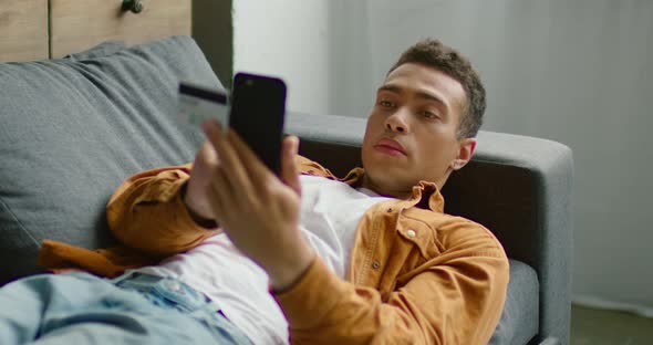 Hispanic Handsome Man Is Lying on Sofa, Holding Smartphone and Credit Card