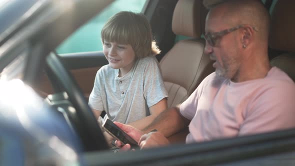 Happy Caucasian Boy Sitting in Car with Man Talking Surfing Internet on Smartphone