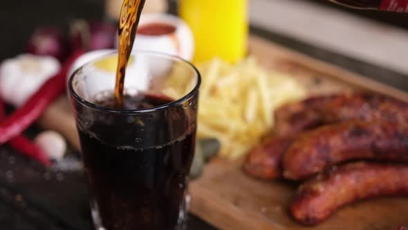 Pouring Soda Drink Into Glass with Tasty Grilled Sausages on Background