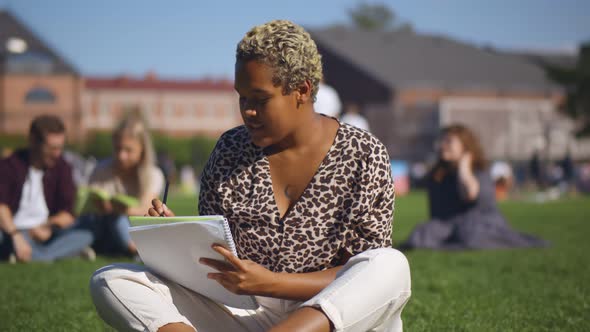 Young African-american Student Woman Writing Notes Sitting on Grass at University Campus