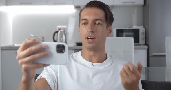 Smiling Young Man Holding Smartphone in Horizontal Position and Making Video Call While Sitting on