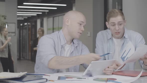 Confident Caucasian Men Discussing Project Plan Sitting at the Table As Blurred Women Talking at the