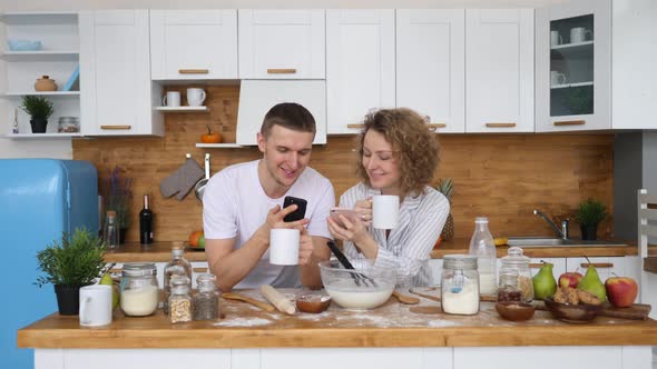 Cute Cheerful Couple Using Smartphone Together At Home In Kitchen
