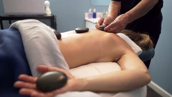 Masseuse or massage therapist uses heated stones on mature, beautiful woman’s back and hands. Steadi