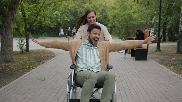 Dolly Shot of Woman Running and Pushing Wheelchair While Disabled Guy Enjoying Fast Ride Smiling in