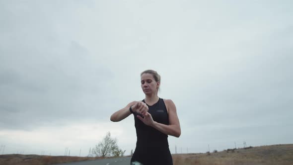 Woman of Athletic Build is Jogging Outdoors and Checks Pulse on Fitness Bracelet Front View