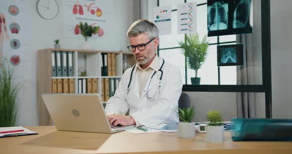 Grey-Headed Doctor Sitting at Workplace in Medical Cabinet and Working on Computer