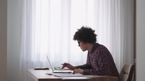 Curly Haired Male Student Attractive Young Boy in Glasses is Studying at Home Using Laptop Typing