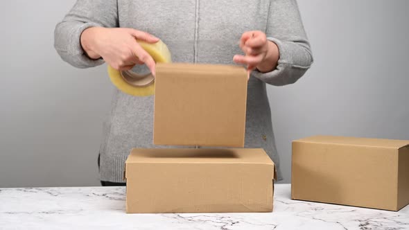 woman in a gray sweater holds a roll of duct tape and packs brown cardboard boxes 