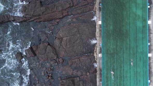 People exercising in an unique coastal swimming pool with waves crashing around. High drone view.