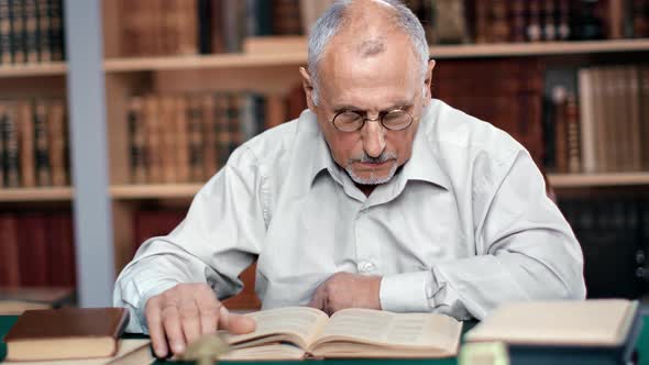 Pensive 70s Grandfather Reading Vintage Paper Text Book Sitting at Desk Table in Public Library